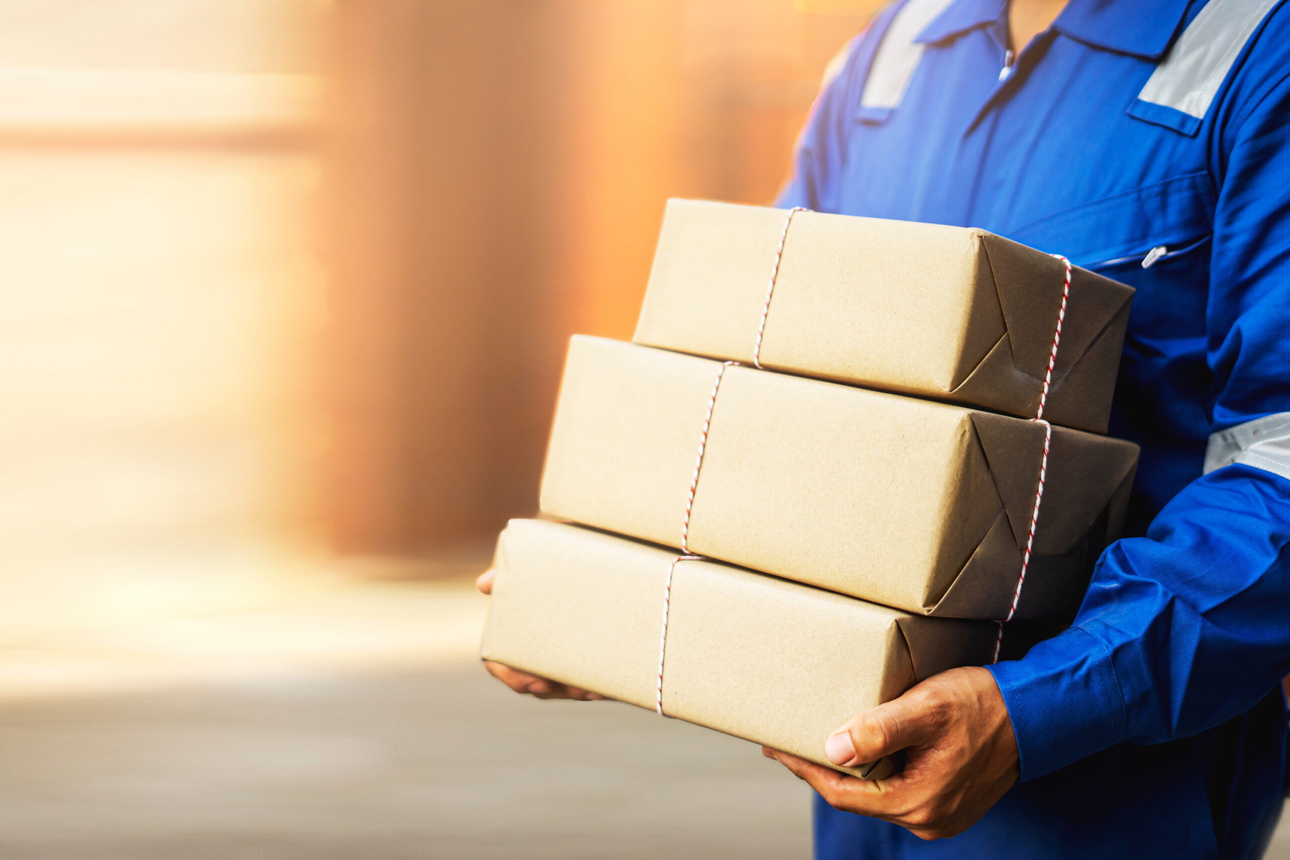 close up asian post delivery man holding boxes of brown parcel with blur background and sunset light, formally wearing blue overall uniform with a hat walking on a delivery through front gate of house